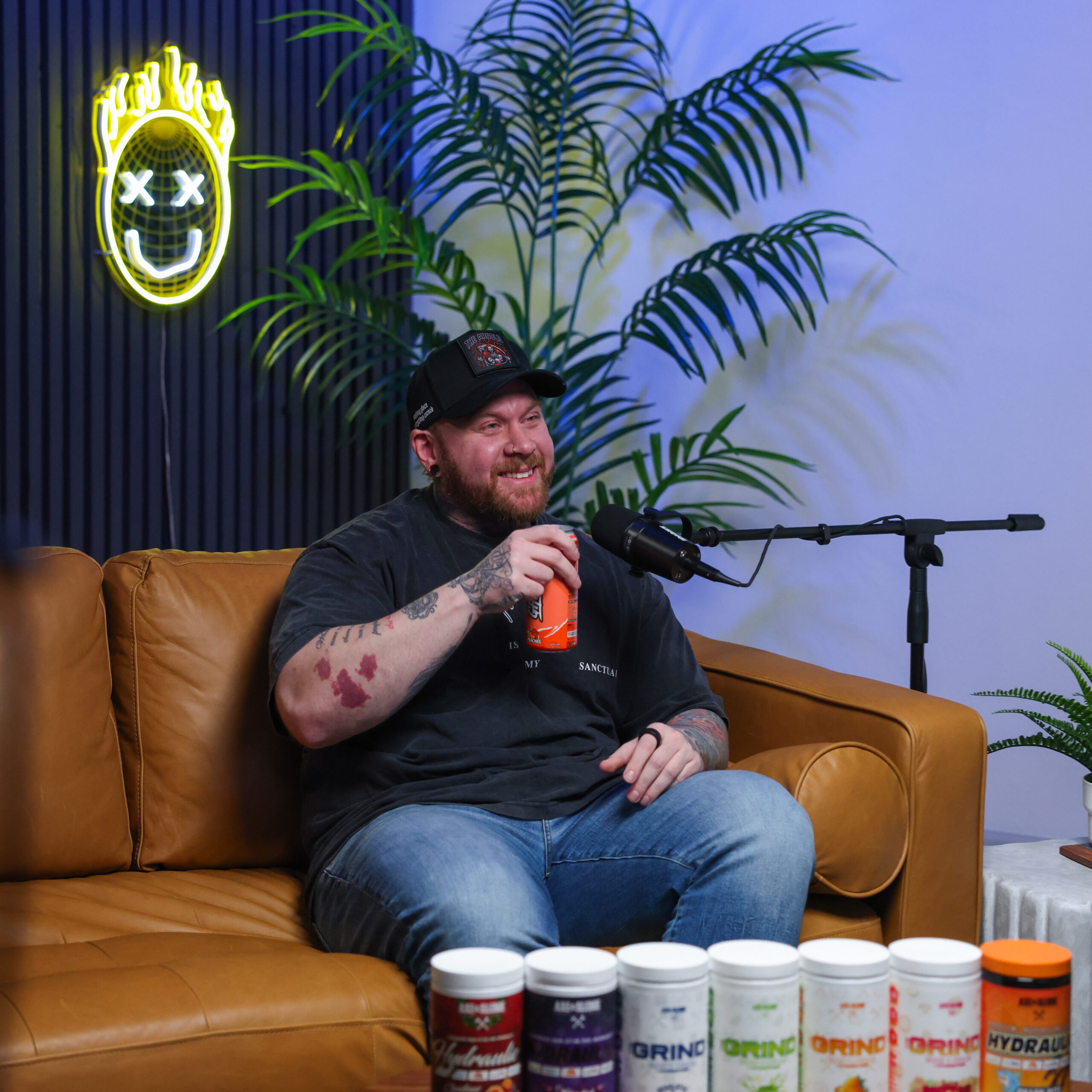 Alt Text: Smiling podcast guest holding a can while sitting on a couch with a display of supplement containers. Visual emphasis on product authenticity and user connection.