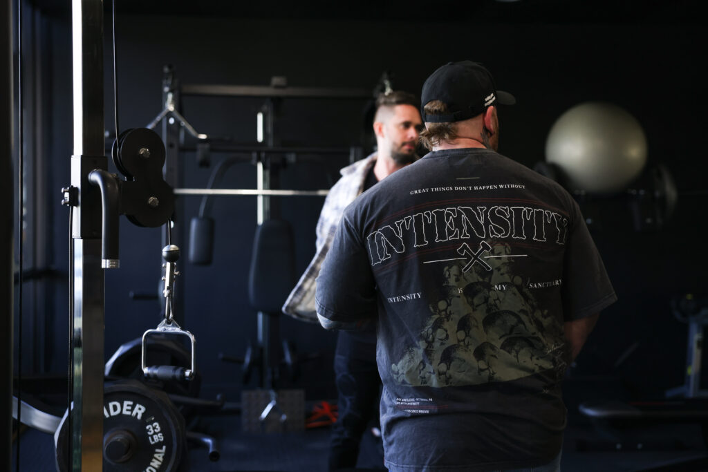 Two men talking inside a gym, one wearing a shirt that says “Intensity.” Fitness-focused setting highlighting brand personality and athlete culture.