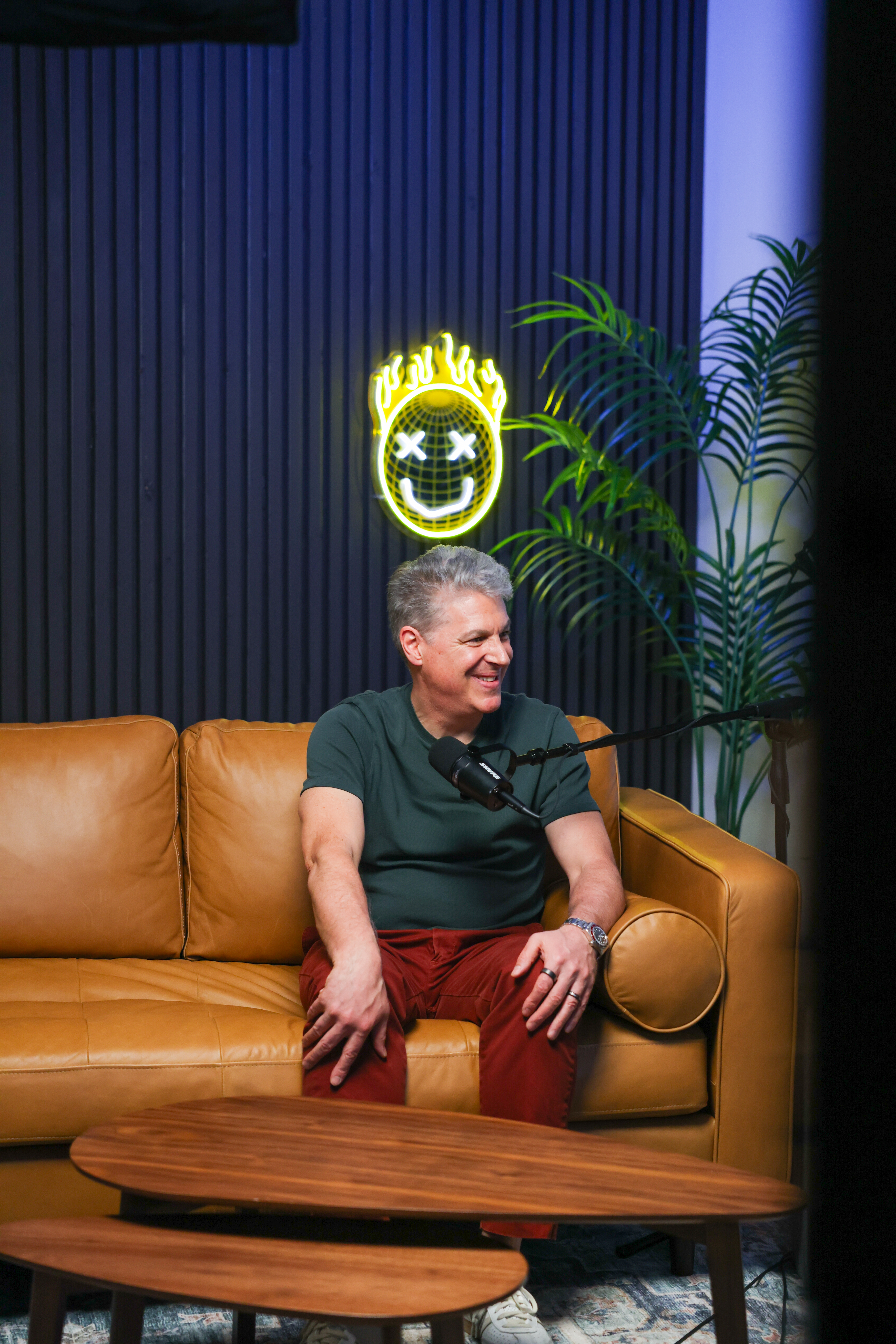 Kenn Israel smiling during a podcast discussion, seated in a modern studio with neon sign and indoor plant.